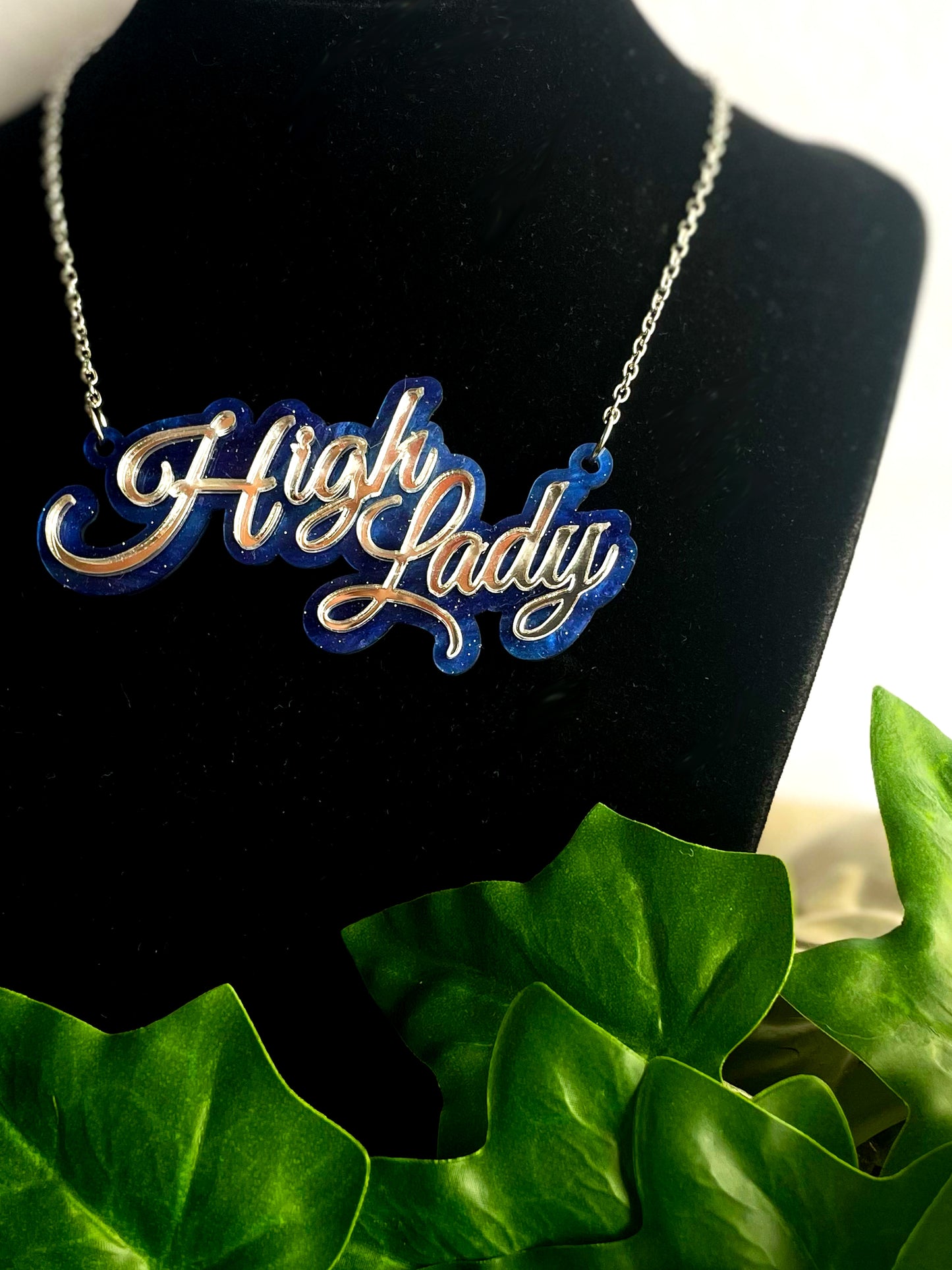 Necklace with 'High Lady' pendant on a black stand with green leaves in the background