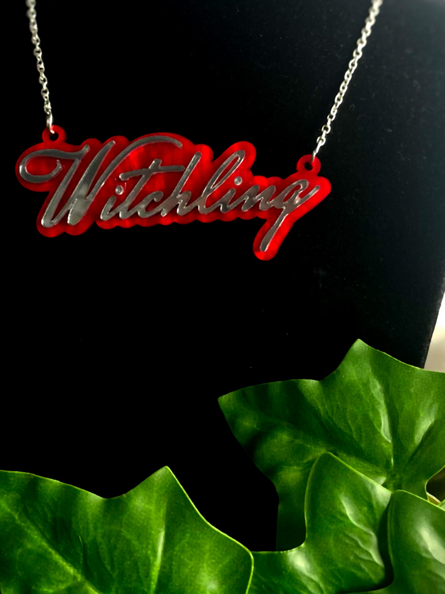 Red 'Witchling' necklace on a black stand with green leaves in the foreground